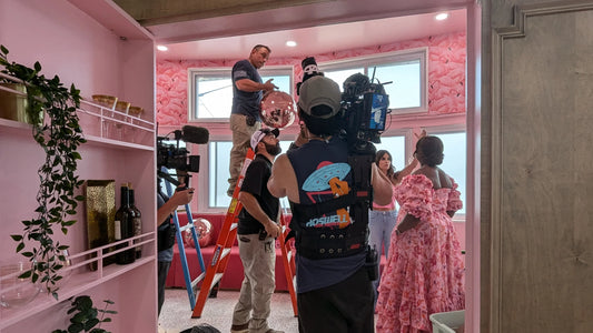 A television crew captures a worker on a ladder hanging a pink disco ball in a vibrant speakeasy lined with pink wallpaper and shelves, while two women in pink outfits look on.